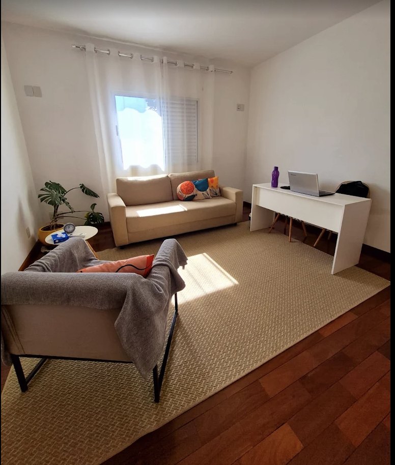Consultório com sofá, poltrona, tapete e escrivaninha, iluminado por luz natural
